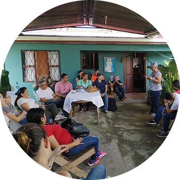 varias personas reunidas en el corredor de una casa escuchando a una señora hablar.