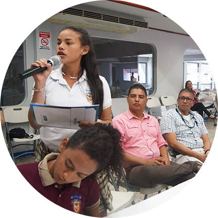 dos mujeres, una sentada y otra de pie hablando con un micrófono, hay dos hombres sentados observándolas.