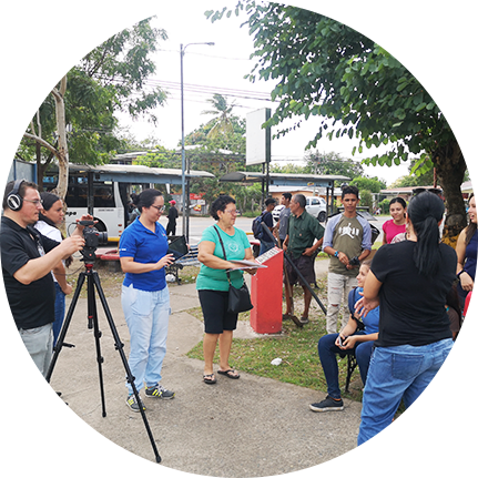 hombre con cámara y trípode, grabando a una mujer sentada en un parque con personas al fondo