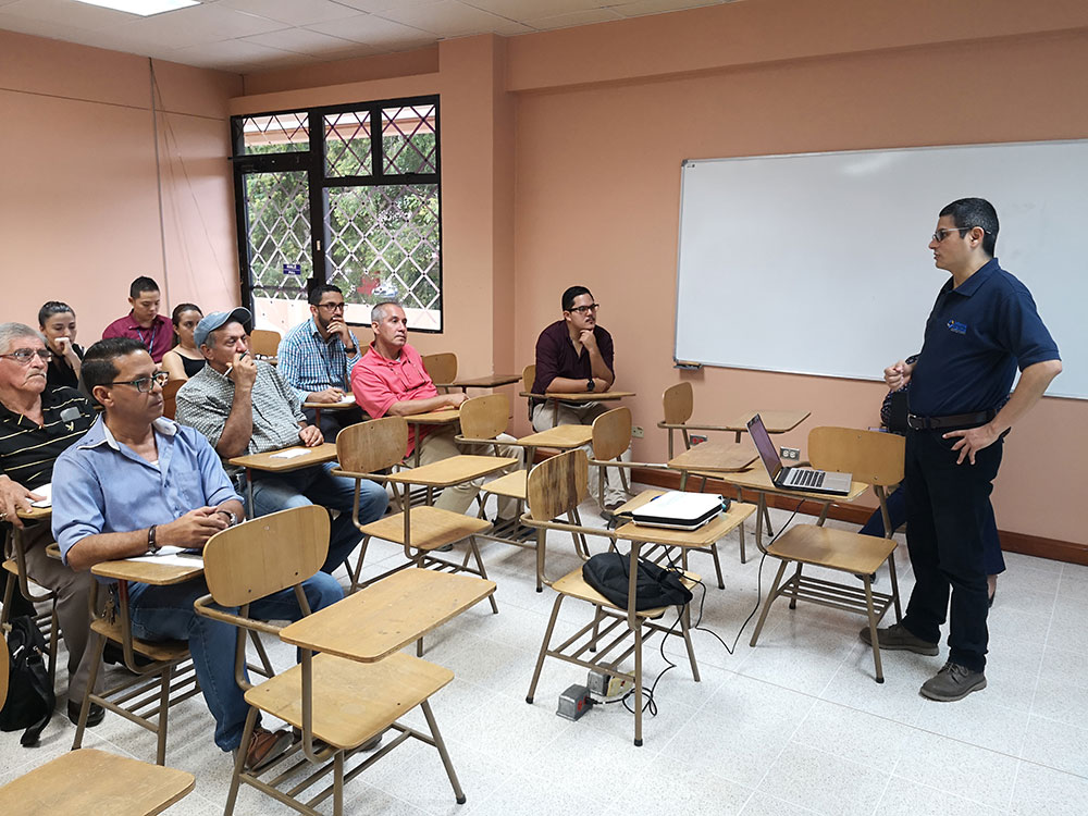 personas en un salón de clases, sentadas en los pupitres viendo al profesor