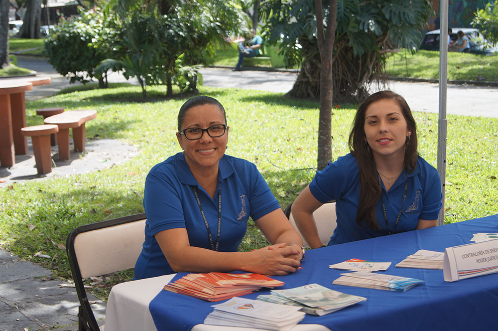 dos mujeres sentadas, sonriendo, con sus manos apoyadas en una mesa que tiene información.
