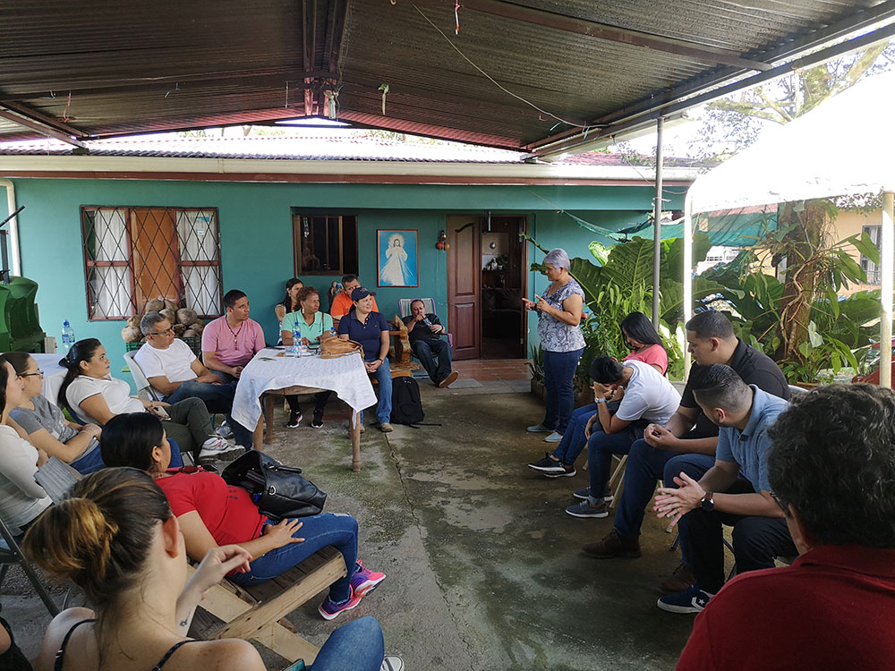 personas sentadas en el corredor de una casa, escuchando a una señora hablar.
